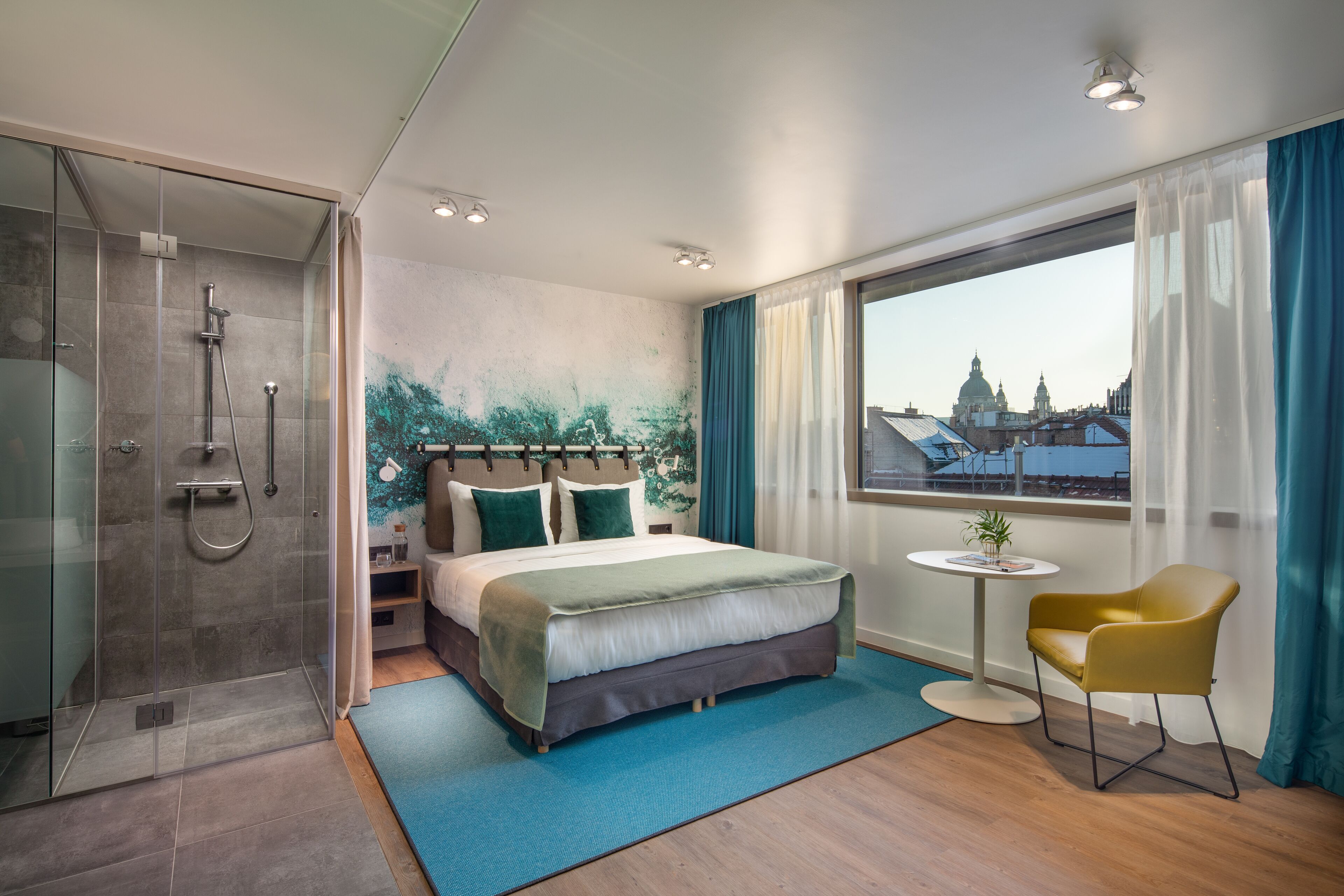 Habitación de hotel moderna con una cama grande, ducha, una mesita con una planta y vistas a la ciudad.