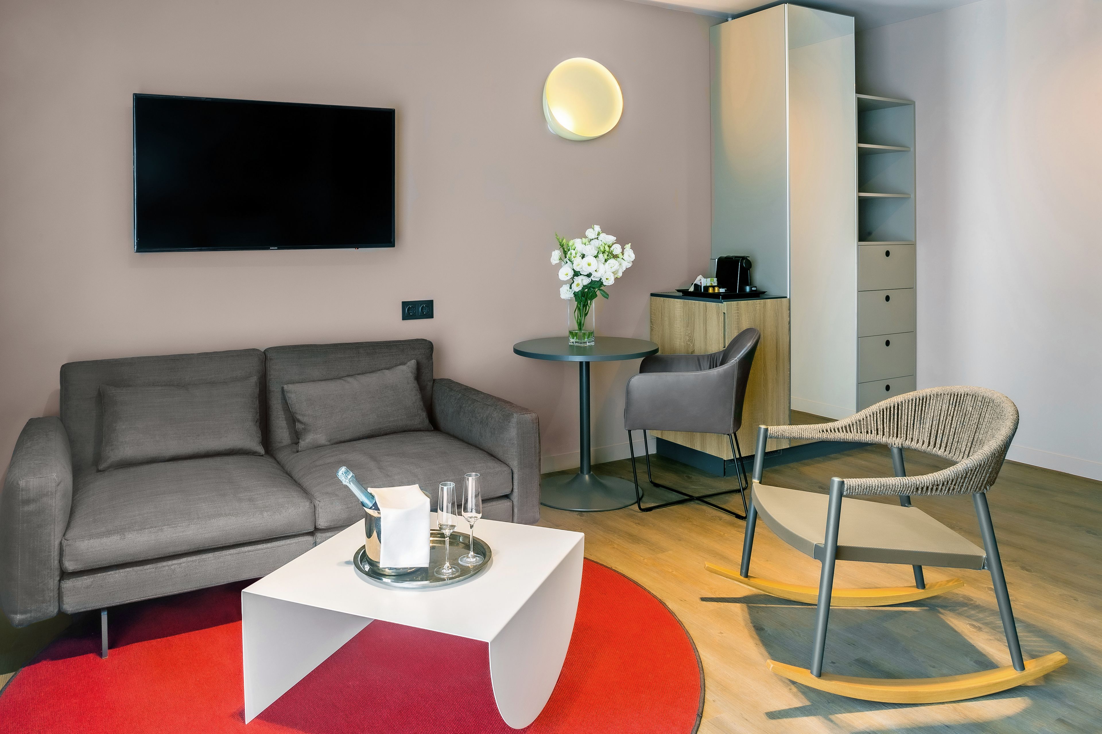 Una sala de estar moderna con un sofá gris, una mesa de centro blanca, un tapete rojo, una mesa redonda con un jarrón de flores, una silla gris y un televisor montado en la pared.
