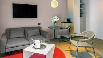 Una sala de estar moderna con un sofá gris, una mesa de centro blanca, un tapete rojo, una mesa redonda con un jarrón de flores, una silla gris y un televisor montado en la pared.