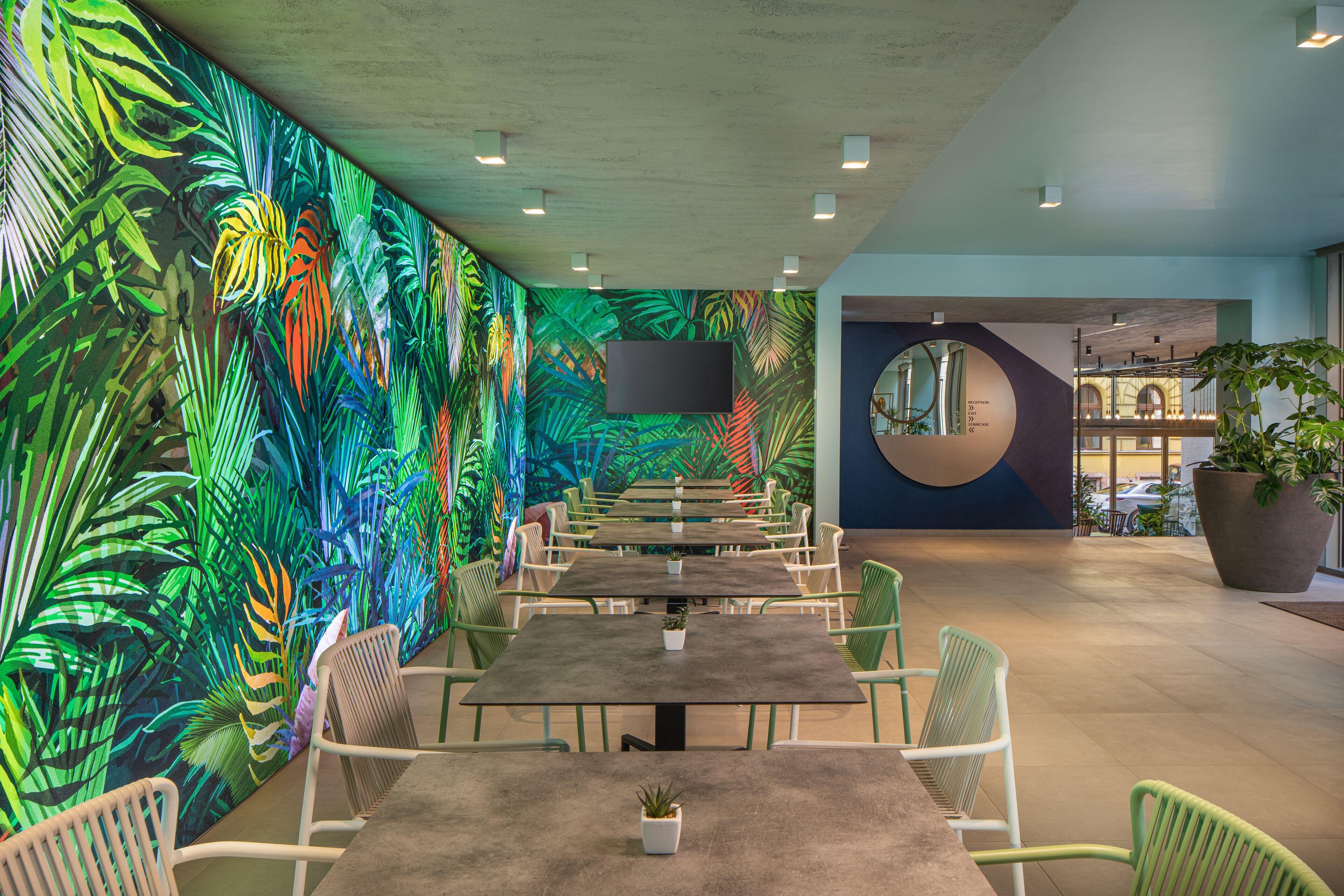 Un espacio interior moderno con una mesa larga y sillas, un gran mural de hojas tropicales y una planta grande en maceta.