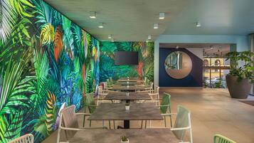 Un espacio interior moderno con una mesa larga y sillas, un gran mural de hojas tropicales y una planta grande en maceta.