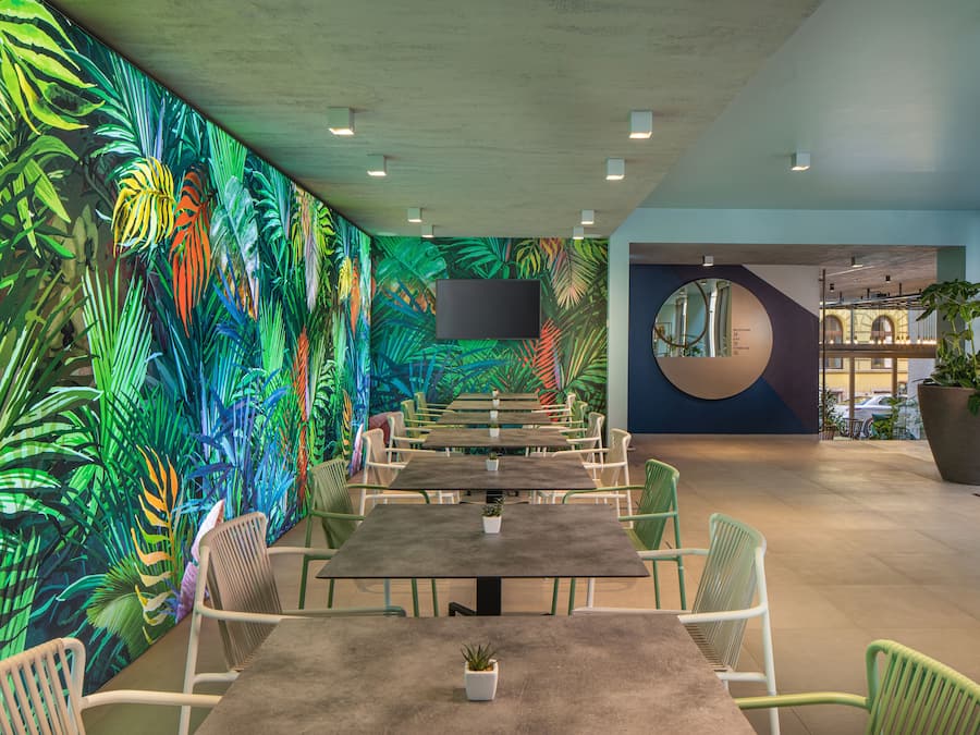 Un espacio interior moderno con una mesa larga y sillas, un gran mural de hojas tropicales y una planta grande en maceta.
