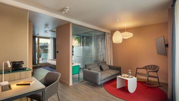 Een moderne hotelkamer met een glazen douche, een slaapbank, een bureau met lamp en een rood vloerkleed.