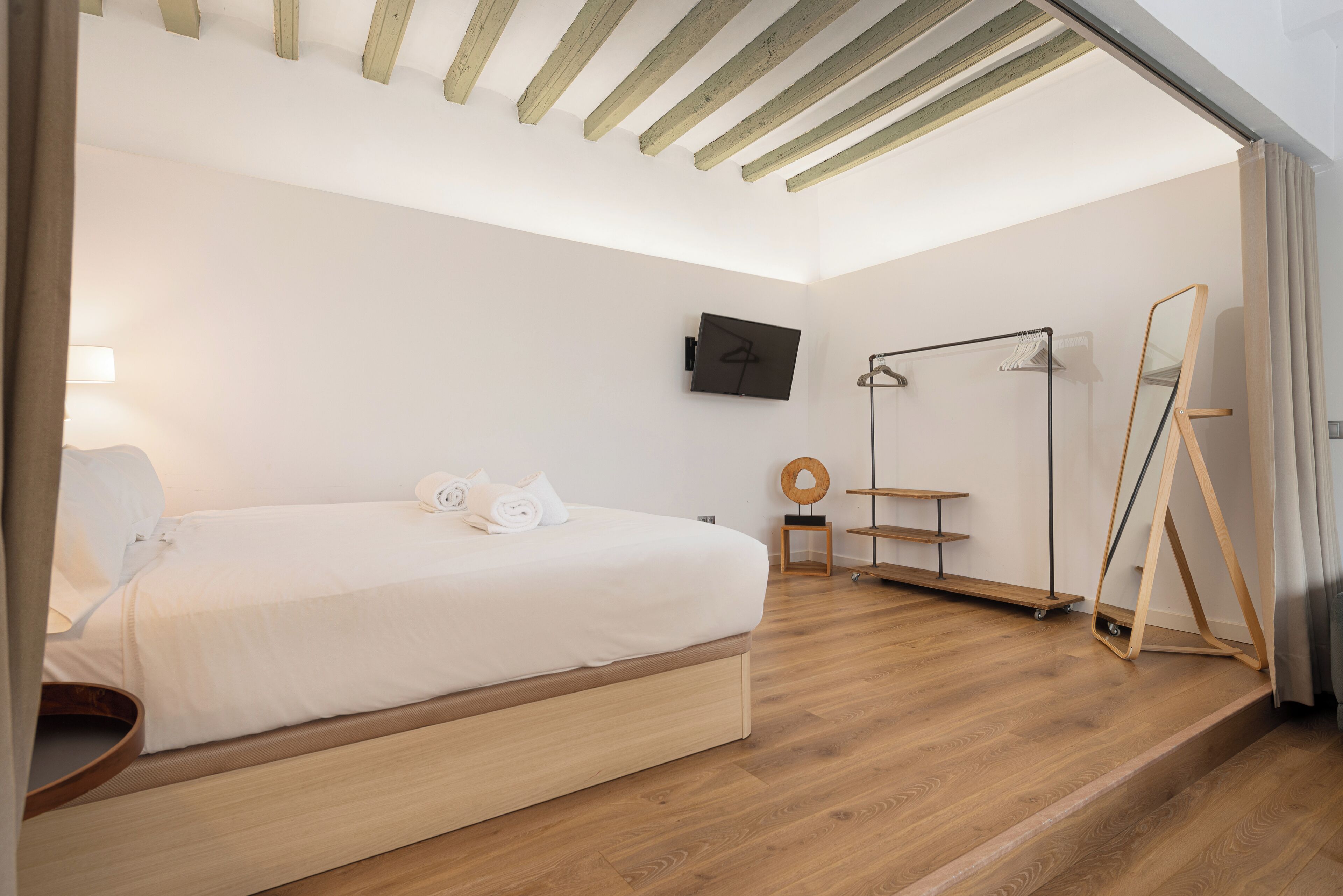 A modern bedroom with a wooden bed, a flat-screen TV, a standing mirror, and a small desk with a chair.