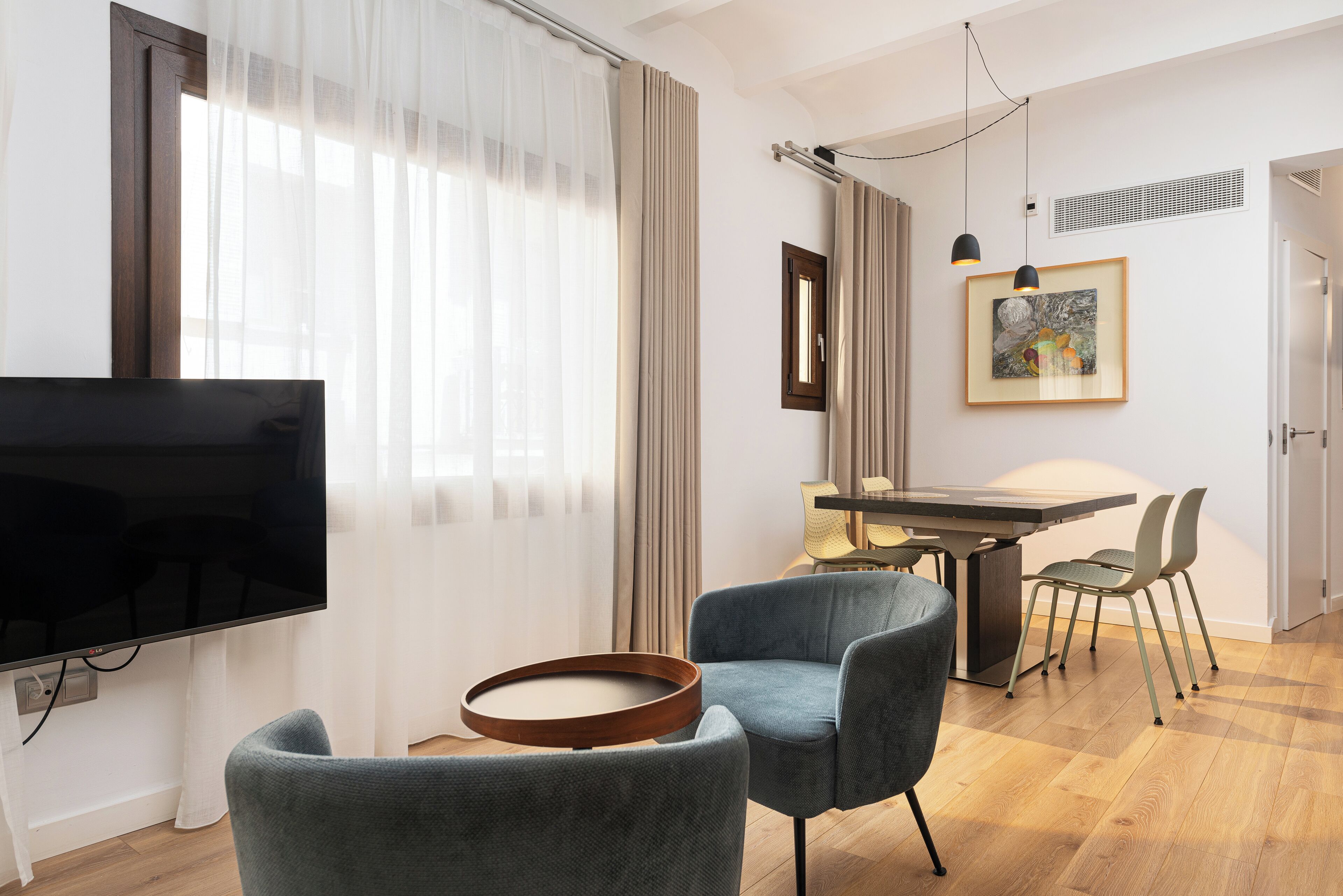 Een moderne woonkamer met een flatscreen-tv, een ronde houten salontafel en twee blauwe fauteuils. Op de achtergrond is een eethoek met een tafel en stoelen te zien.