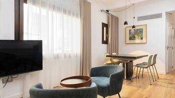 Een moderne woonkamer met een flatscreen-tv, een ronde houten salontafel en twee blauwe fauteuils. Op de achtergrond is een eethoek met een tafel en stoelen te zien.