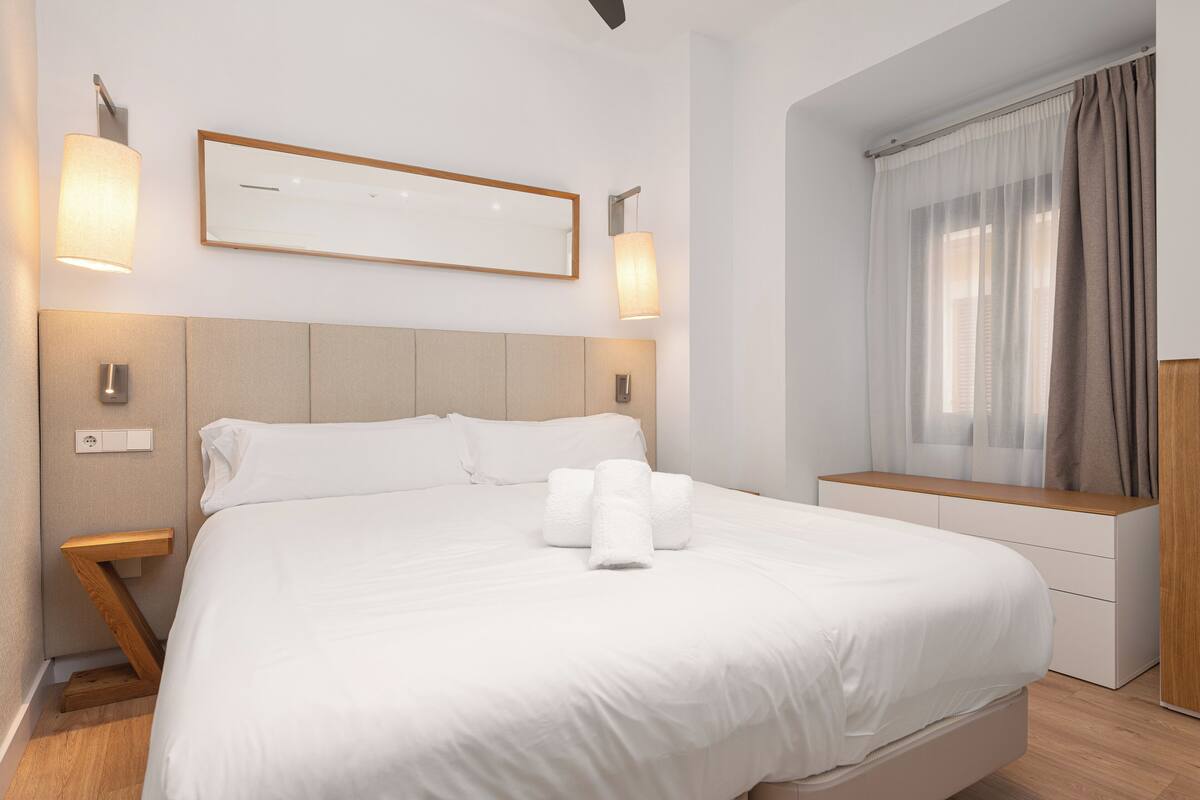 Une chambre d’hôtel moderne dotée d’un grand lit, de tables de chevet, d’une banquette intégrée, d’un miroir et d’une fenêtre avec des rideaux.