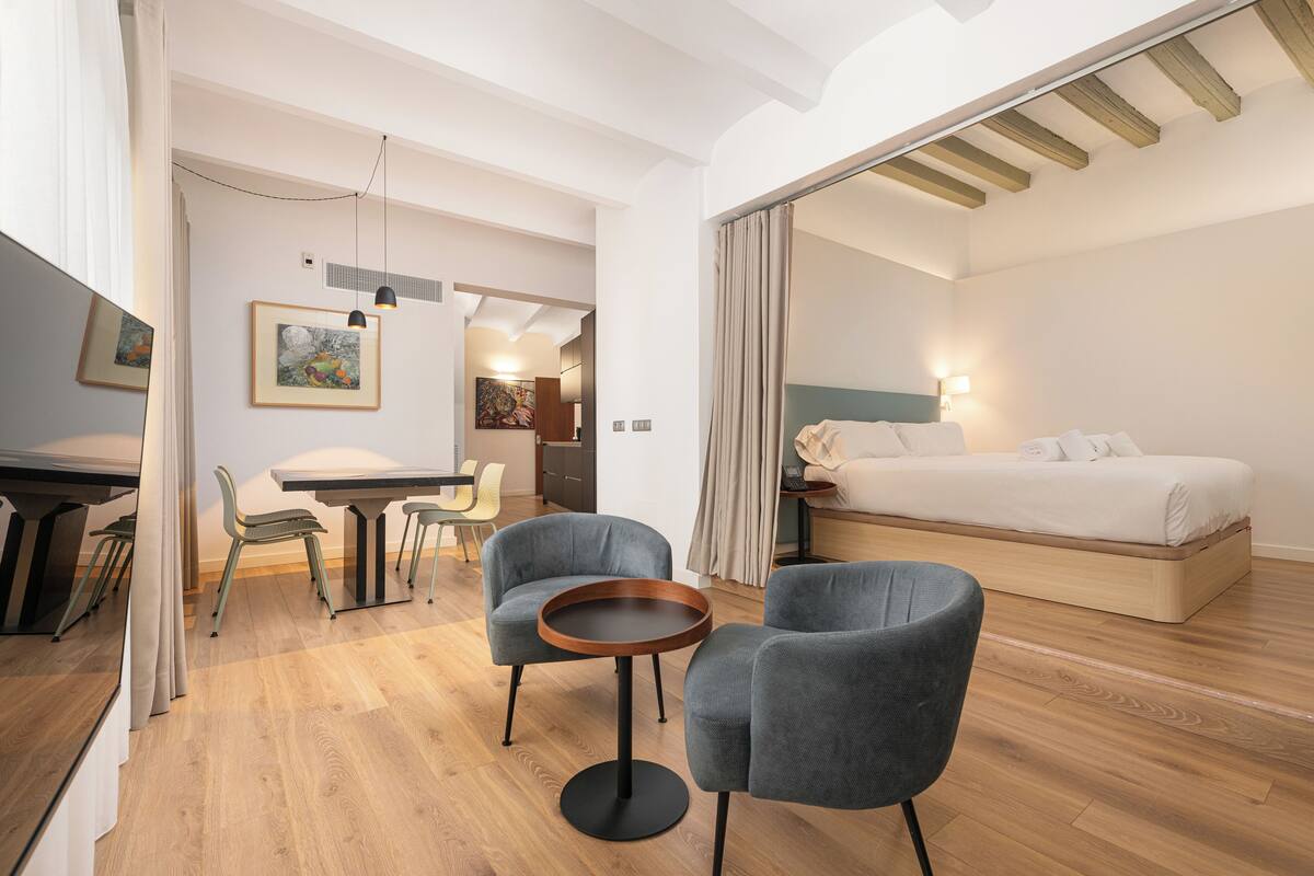 Une chambre d’hôtel moderne avec un plancher en bois, un lit recouvert de draps blancs, une table ronde avec deux chaises et un bureau avec une chaise.