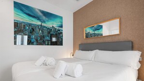 Hotelkamer met een groot bed, witte lakens, twee opgerolde handdoeken, een stadsgezicht schilderij en een spiegel aan de muur.