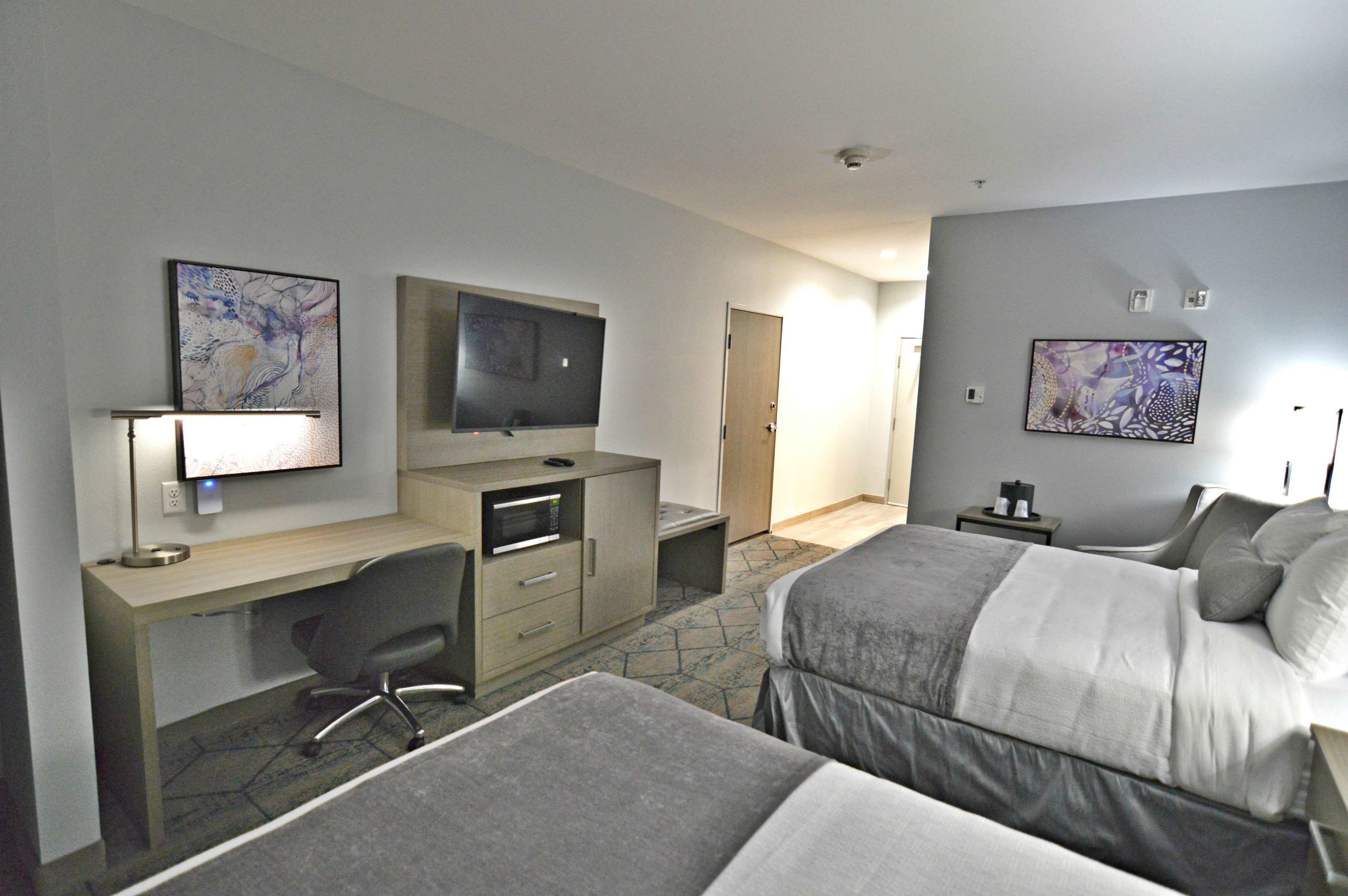 Una habitación de hotel con dos camas, un escritorio con computadora, un televisor y obras de arte en las paredes.