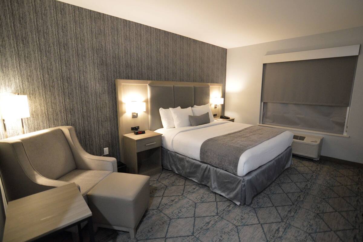 A hotel room with a large bed, a chair, a nightstand with a lamp, and a window with a blind.