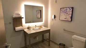 A modern bathroom with a wooden vanity, a large mirror, a framed artwork, a handrail, and a toilet.