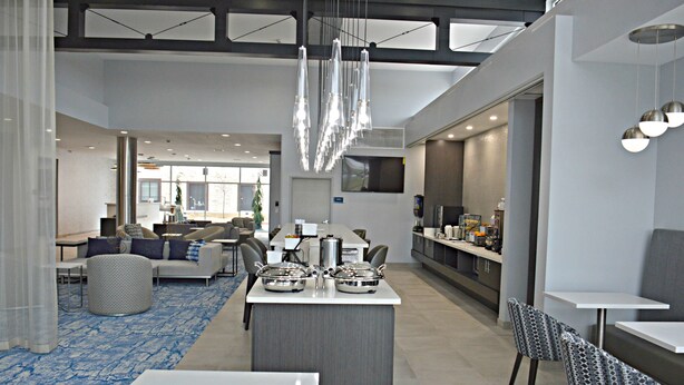 A modern hotel lobby with a central island, seating areas, and a kitchen in the background.