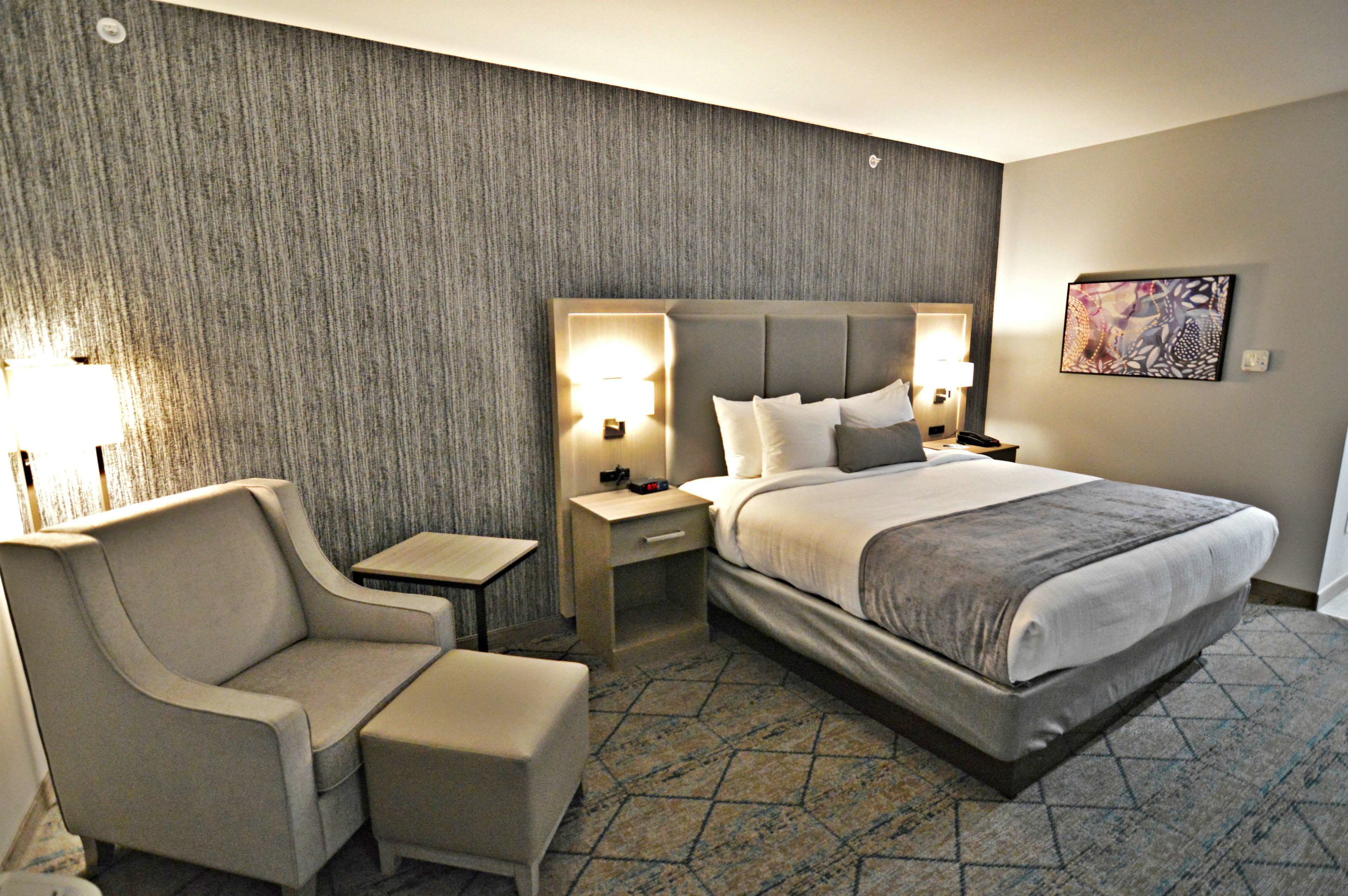 Habitación de hotel con una cama grande, dos mesitas de noche con lámparas, una silla y suelo alfombrado.