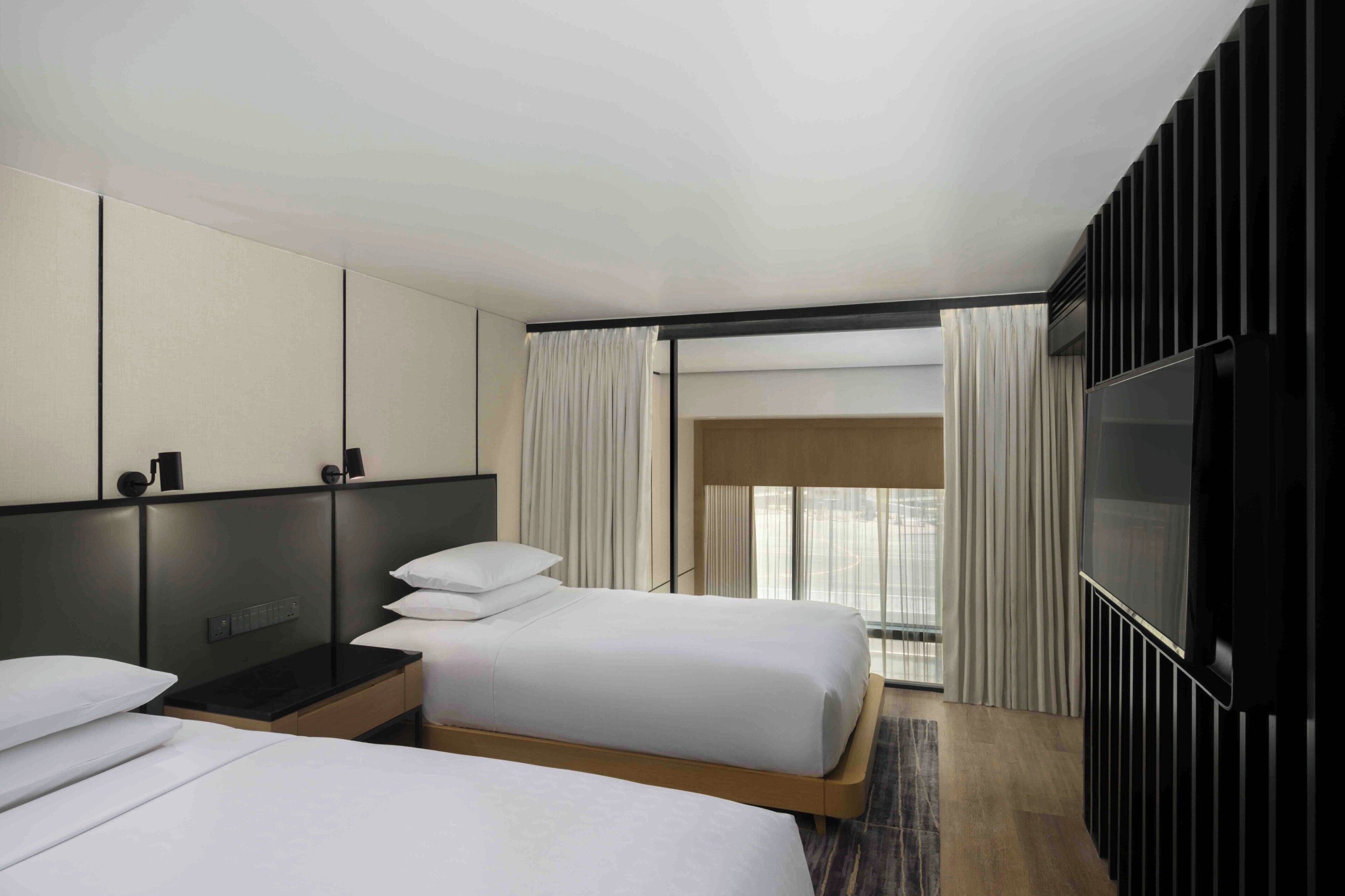 A hotel room with two beds, a TV, a desk, and a large window with curtains.