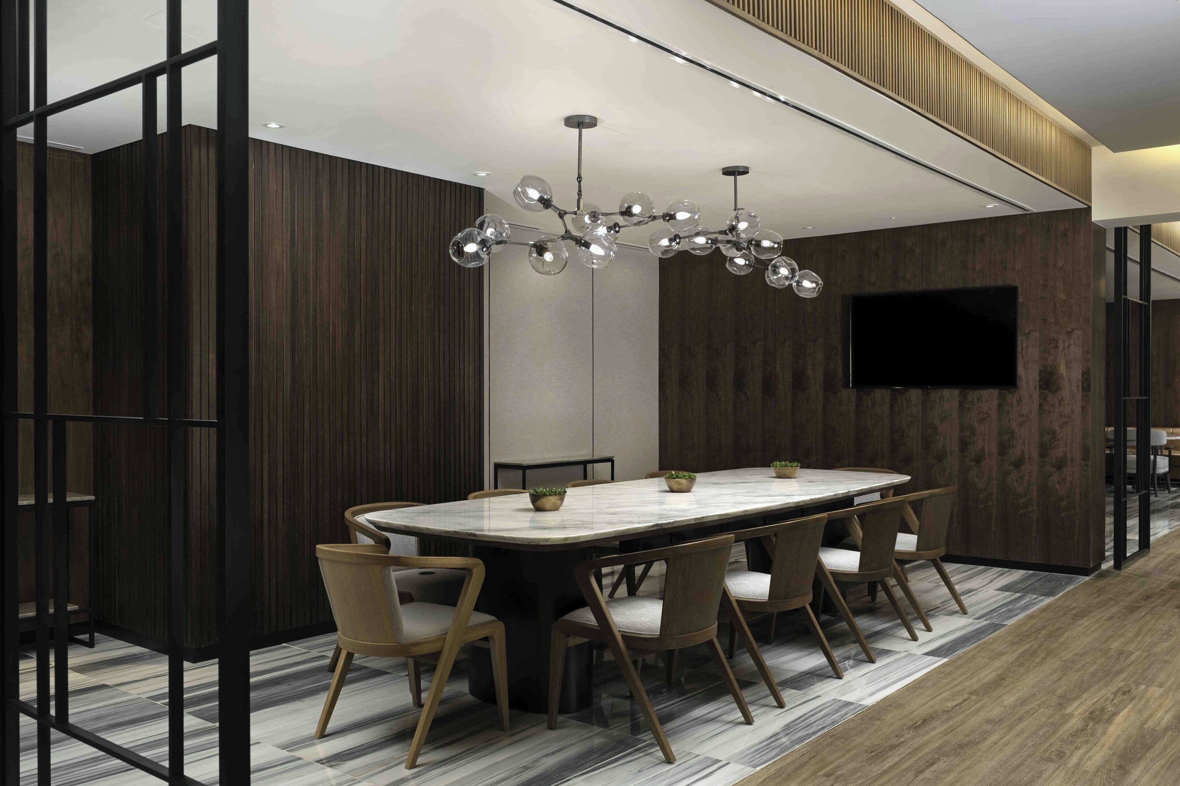 Un espace repas moderne avec une grande table rectangulaire, six chaises, un lustre original et un téléviseur à écran plat fixé au mur.