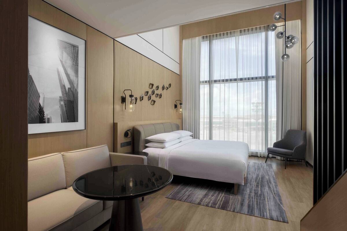 A modern hotel room with a bed, a sofa, a small table, and a large window with curtains.