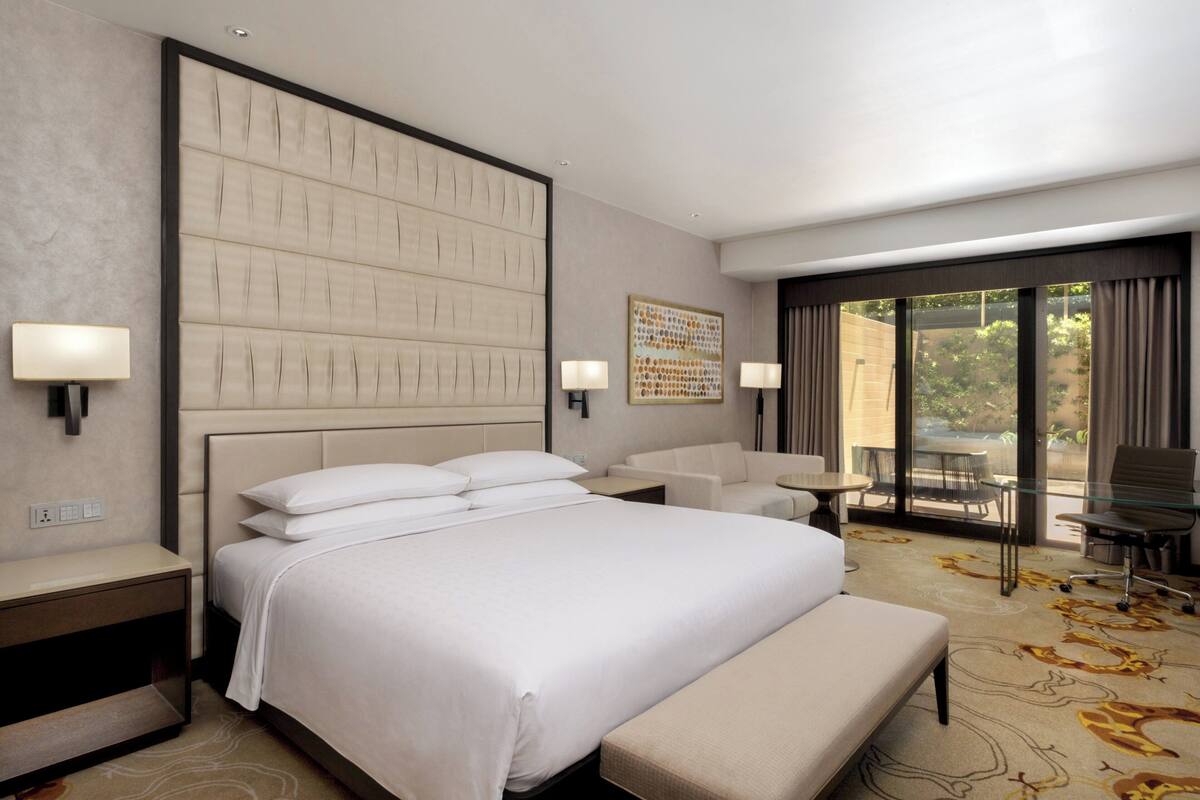 A hotel room with a large bed, a bench, two bedside tables, a desk, and a chair. There is a large window with a view of greenery outside.