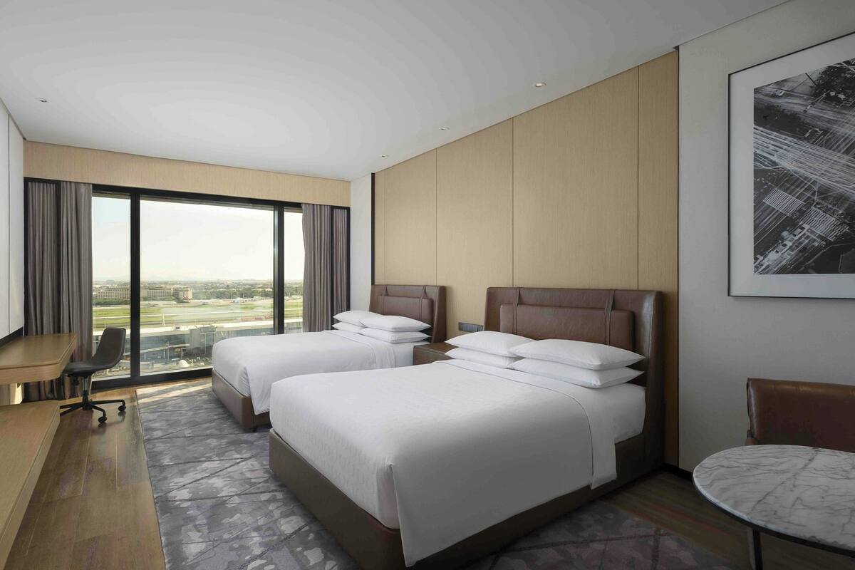 A hotel room with two beds, a desk, a chair, and a view of the cityscape.
