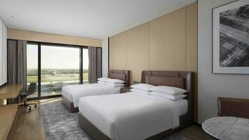 A hotel room with two beds, a desk, a chair, and a view of the cityscape.