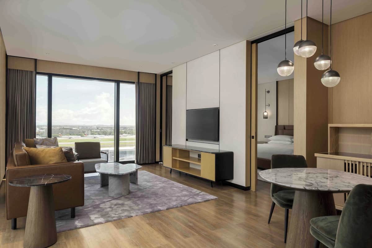 A modern hotel room with a large window, a flat-screen TV, a sofa, a coffee table, a dining table, and a small kitchen area.