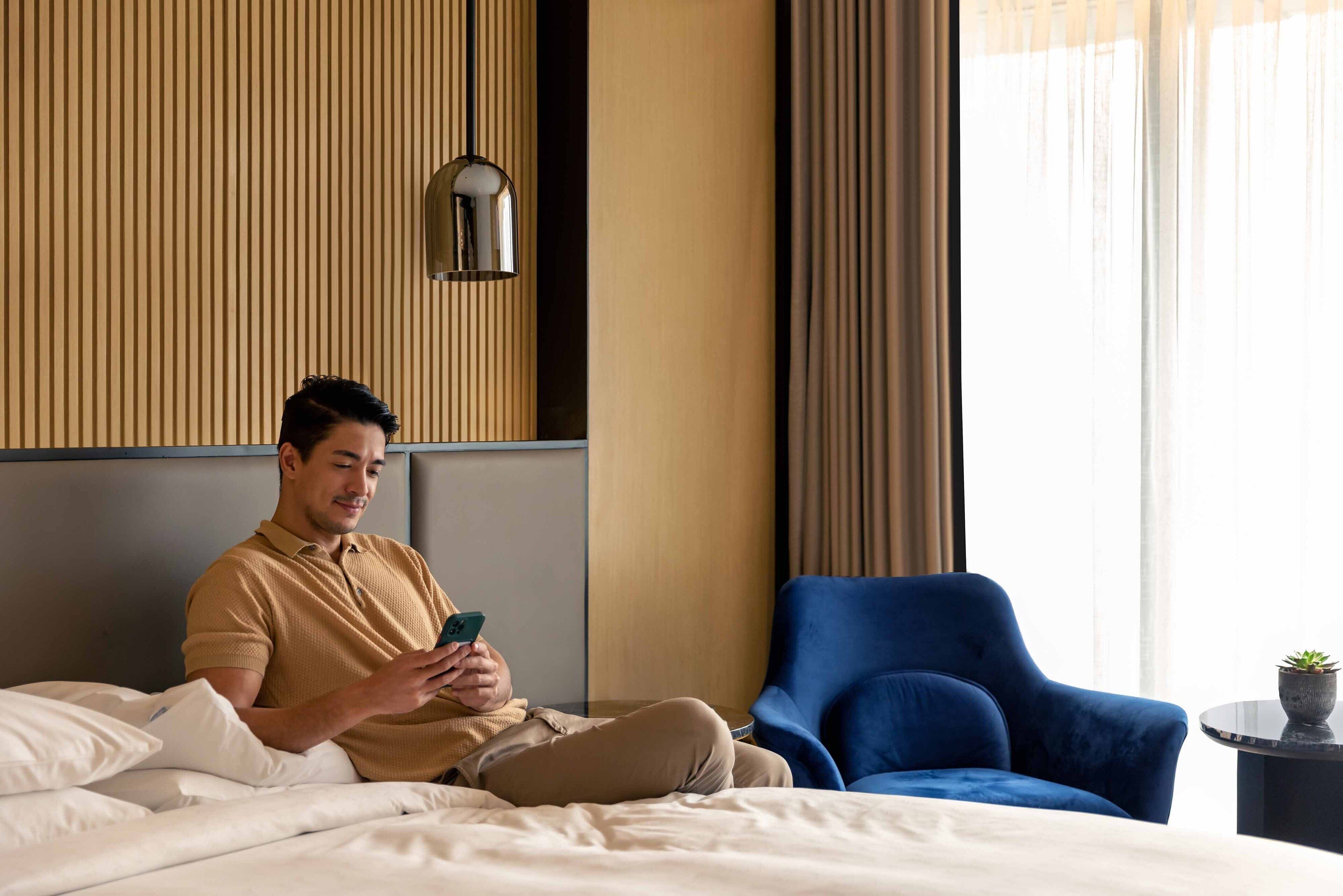 Een man zit op een bed in een hotelkamer en gebruikt een smartphone.