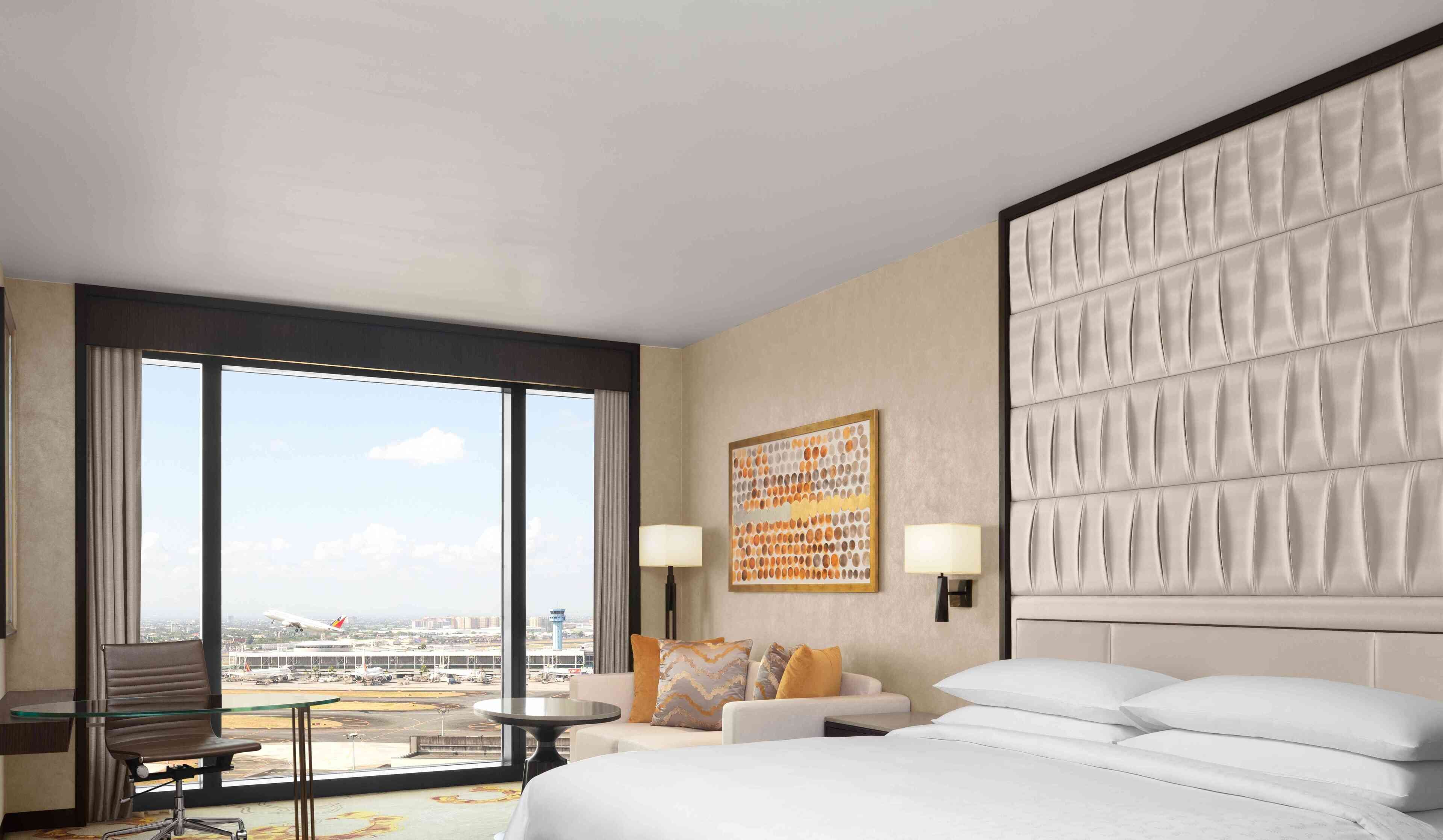 A hotel room with a large bed, a sofa, a desk, and a view of an airport.