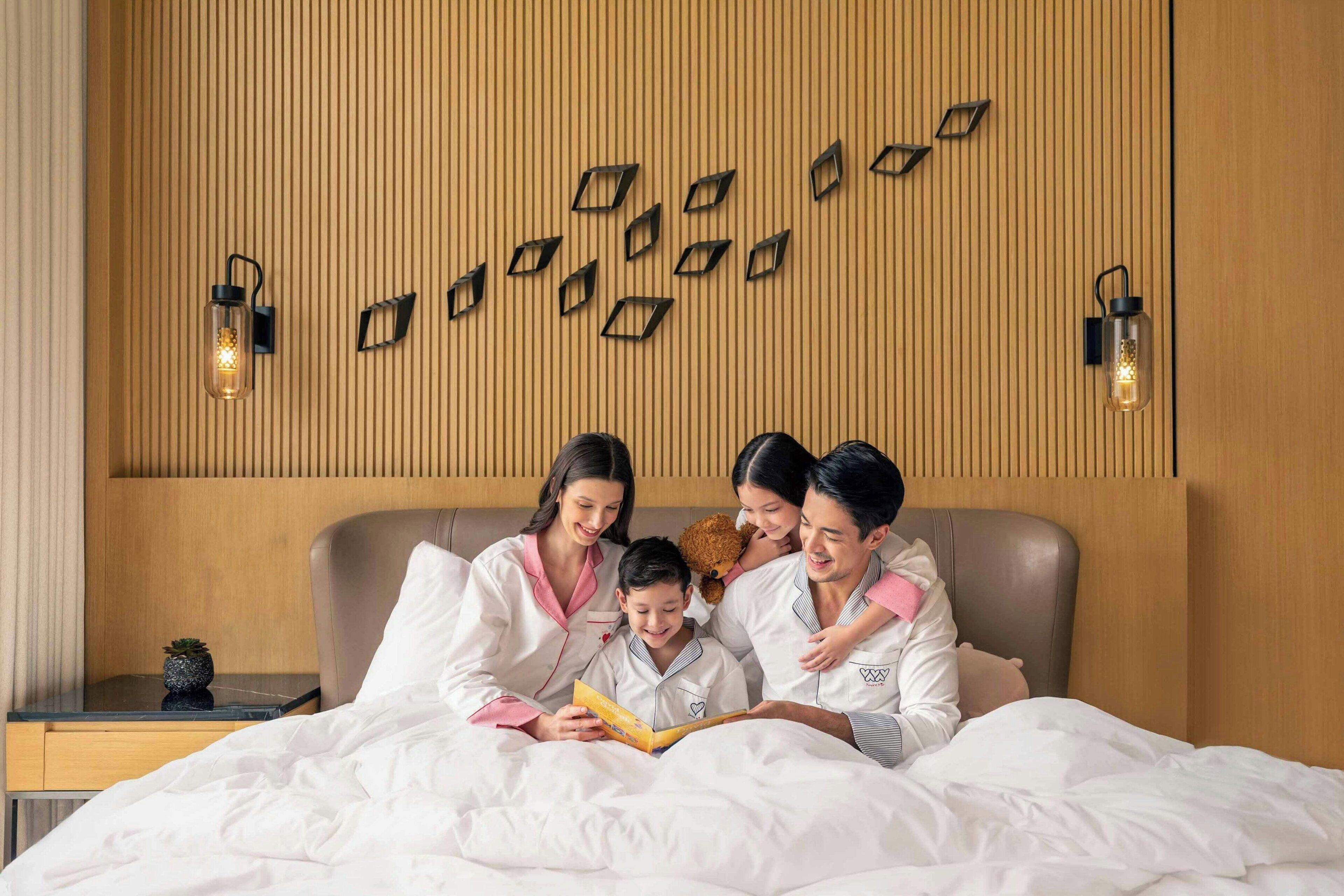 Une famille de quatre personnes se trouve dans une chambre d’hôtel. On y voit un lit en bois avec une tête de lit, une table de chevet avec une lampe et une petite plante en pot.