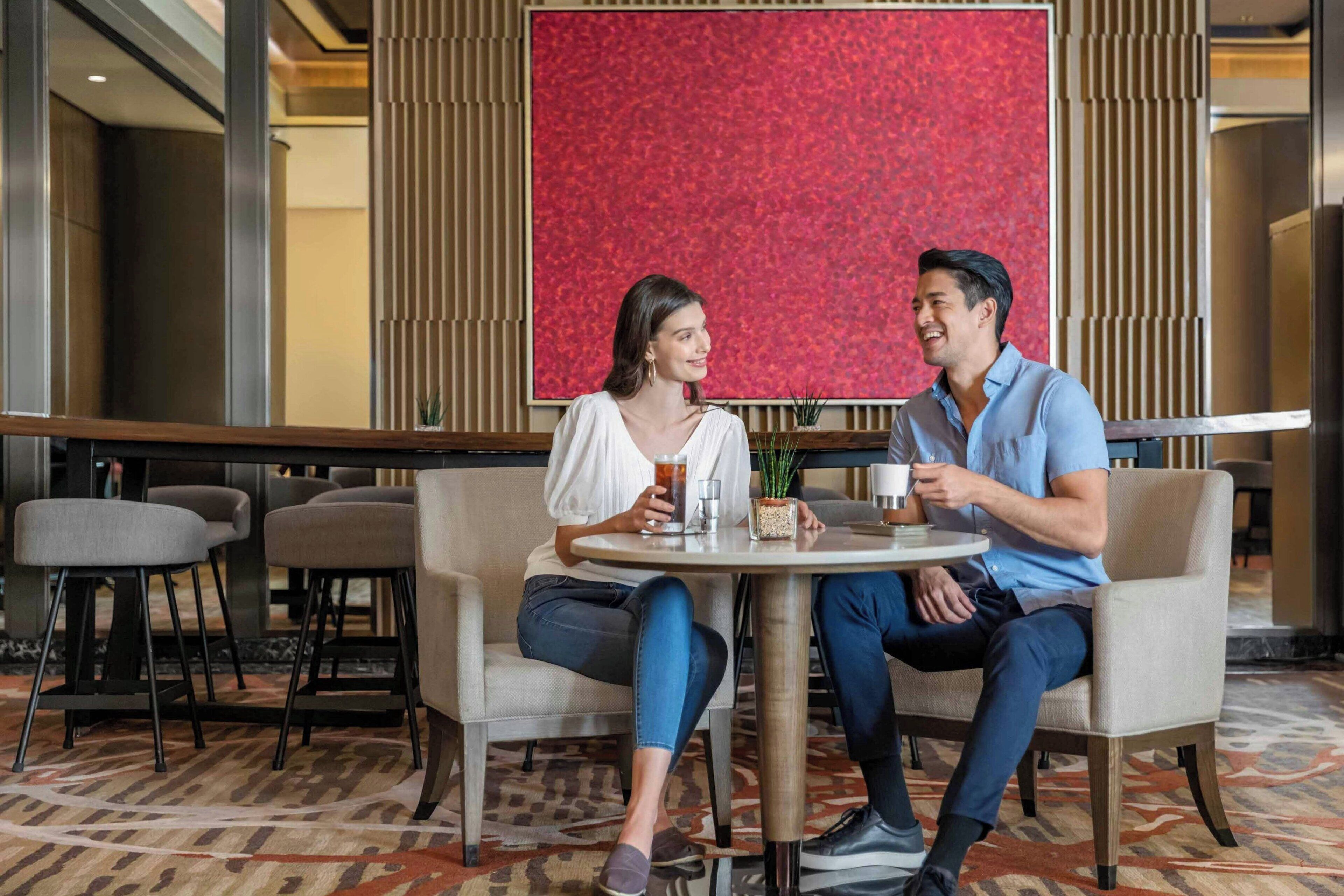 A couple seated at a round table in a modern indoor setting with a red textured wall and patterned carpet.