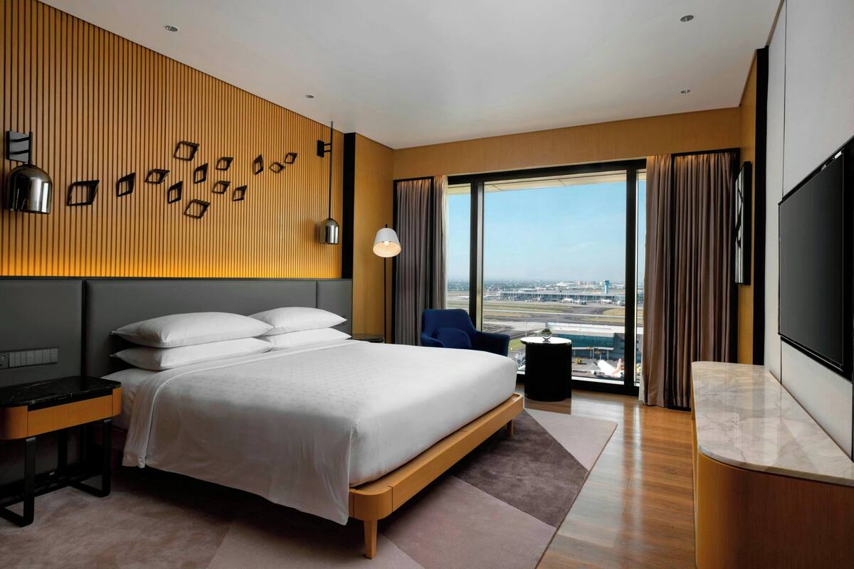 A modern hotel room with a large bed, a desk, a TV, and a view of the city through large windows.