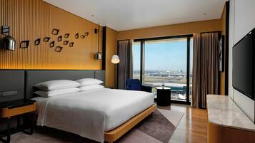 A modern hotel room with a large bed, a desk, a TV, and a view of the city through large windows.