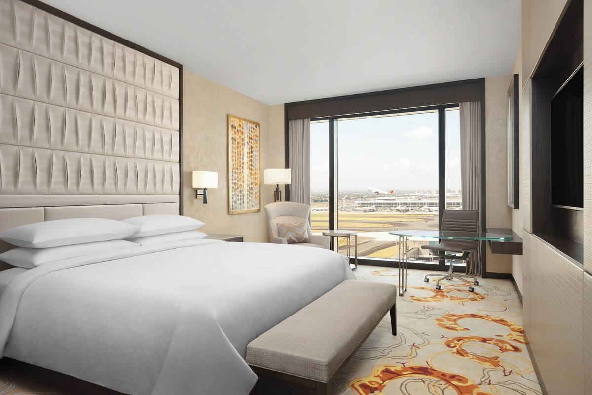 A hotel room with a large bed, a desk with a chair, a view of the city through large windows, and a modern design.