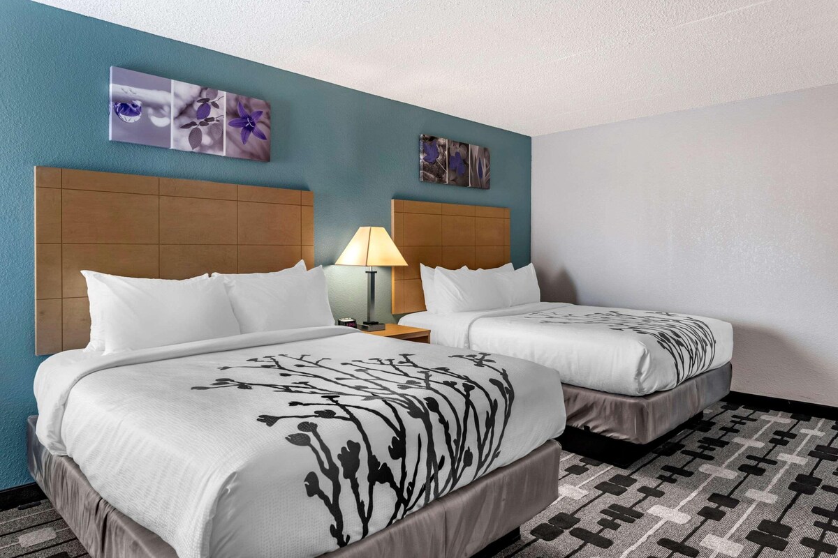 A hotel room with two beds, a headboard, a nightstand with a lamp, and artwork on the wall.