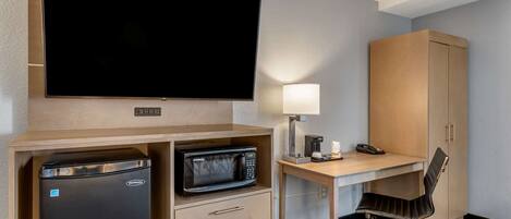 A hotel room with a flat-screen TV, a microwave, a refrigerator, a desk with a lamp, a chair, and a wardrobe.