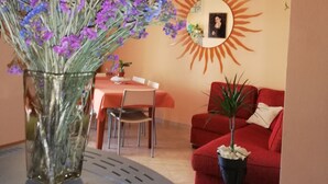 A living room with a red sofa, a dining table with chairs, a vase with flowers, and a wall-mounted sun decoration. - B&B Casa Mia (Alghero)