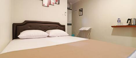 A hotel room with a large bed, a headboard, a wall-mounted artwork, a shelf with a kettle and water bottle, and a visible bathroom door.