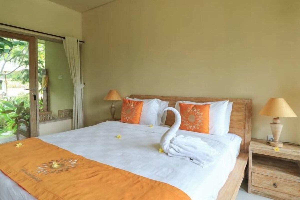 A neatly made bed with a swan-shaped towel, orange and white pillows, and a wooden nightstand with a lamp.