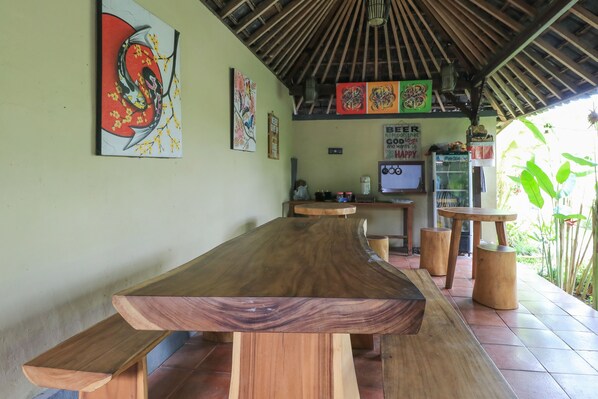 A wooden dining table with benches, surrounded by artwork on the walls, a signboard, and a small refrigerator.