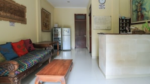 A living room with a sofa, wooden coffee table, refrigerator, and a reception desk with a clock and a sign. - Surfaris Inn on Poppies 2 (Kuta)