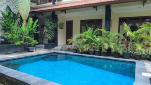 A swimming pool with a tiled edge, surrounded by potted plants and a building with a tiled roof and wooden doors. - Surfaris Inn on Poppies 2 (Kuta)