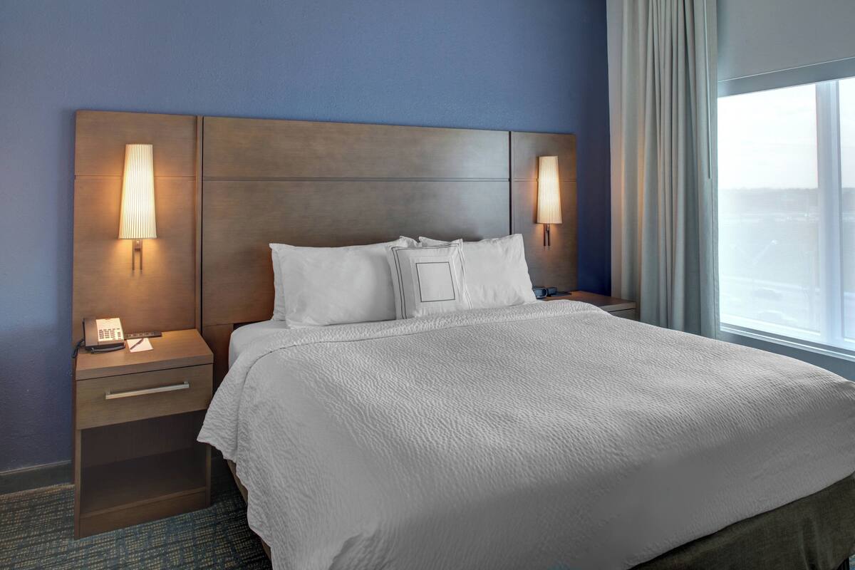 Une chambre d’hôtel avec un grand lit, deux tables de chevet avec des lampes, une petite table de nuit avec un téléphone et une fenêtre avec des rideaux.