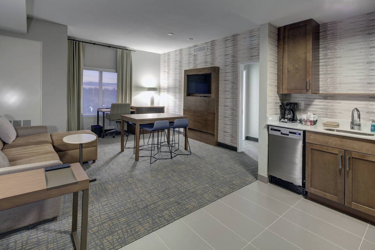 Une chambre d’hôtel moderne équipée d’un canapé, d’une table à manger avec des chaises, d’un téléviseur fixé au mur, d’un coin cuisine avec un lave-vaisselle et d’une grande fenêtre donnant sur un paysage agréable.