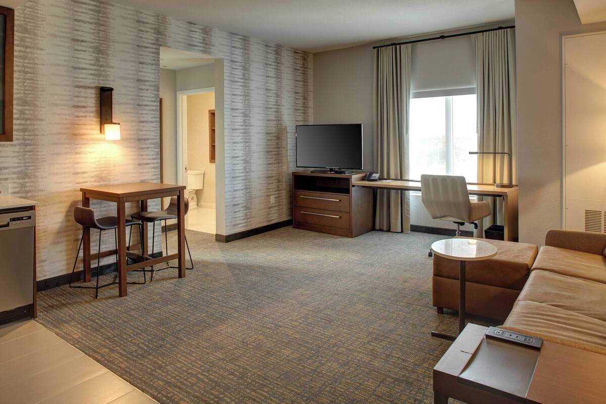 Une chambre d’hôtel équipée d’une télévision à écran plat, d’un bureau avec une chaise, d’un canapé, d’une petite table et d’un mini-réfrigérateur.