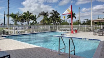 A swimming pool with a flag, palm trees, and a fenced area with chairs and tables.