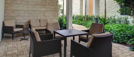 A patio area with wicker furniture, a flat-screen TV mounted on the wall, and a view of a landscaped garden with palm trees and shrubs.