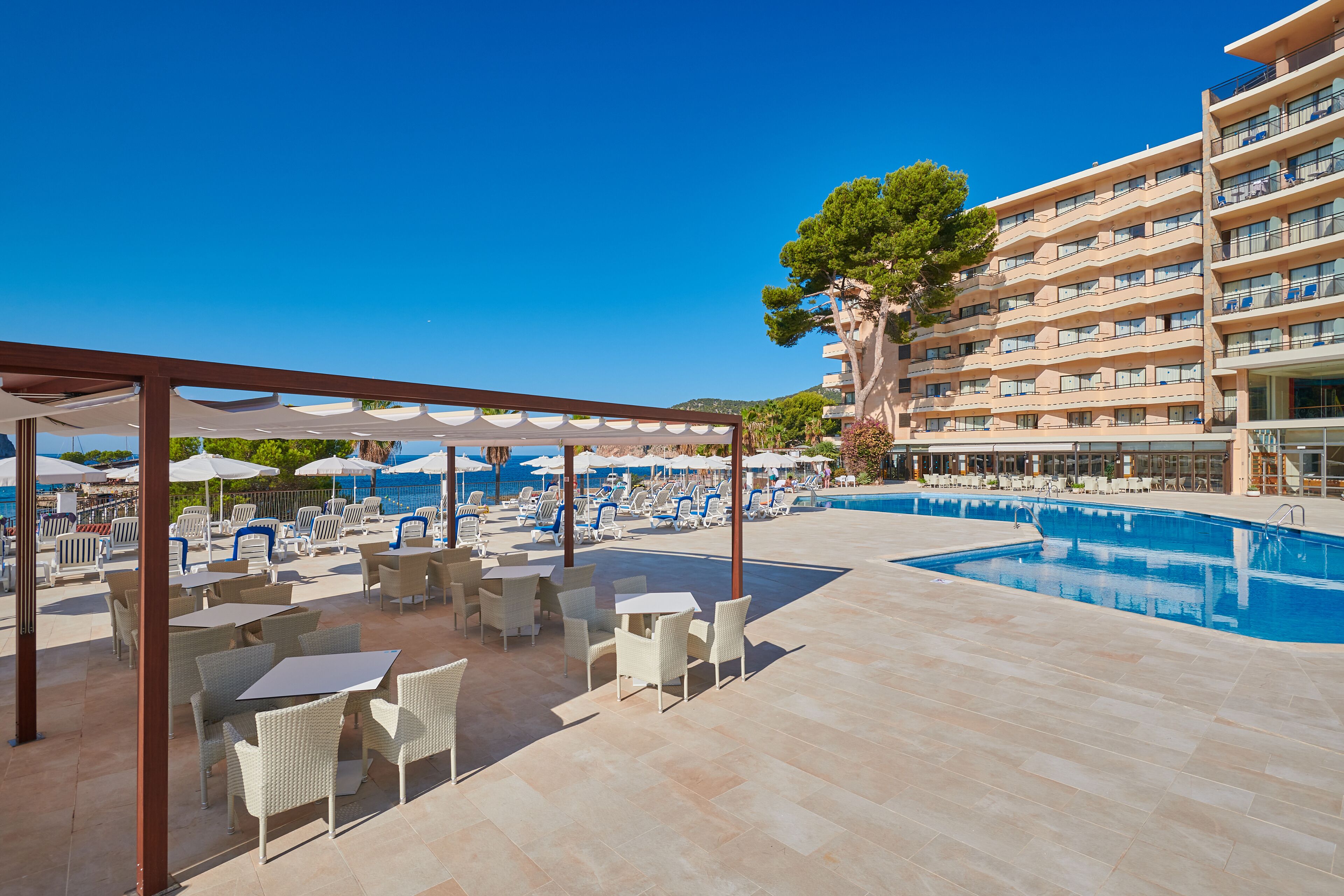 Un espace aménagé au bord de la piscine, avec des chaises longues et des parasols, une piscine, et un bâtiment doté de balcons.