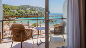 Un balcón con sillas de mimbre y una mesa, con vista a una playa y un paisaje montañoso.
