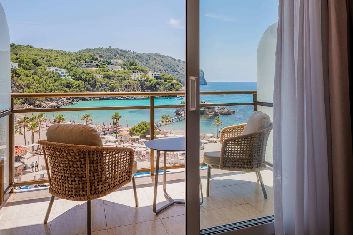 Un balcón con sillas de mimbre y una mesa, con vista a una playa y un paisaje montañoso.