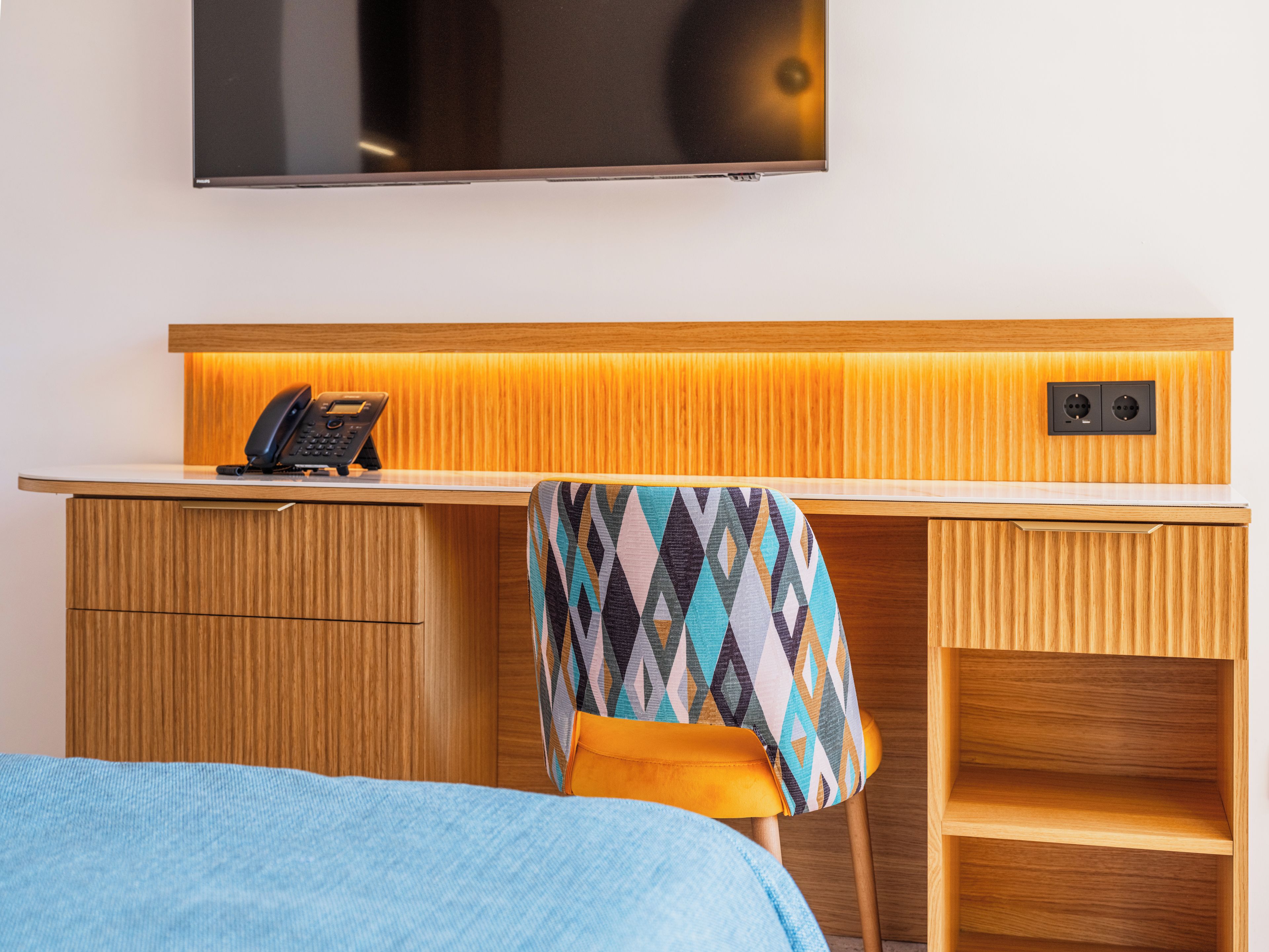 Ein Hotelzimmer mit einem Holztisch, einem Telefon, einem an der Wand befestigten Fernseher und einem gemusterten Sessel.