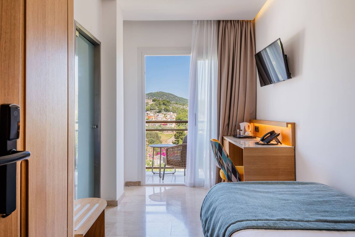 Habitación de hotel con una cama, un escritorio con teléfono, una silla, una mesita y un televisor en la pared. Tiene un balcón con vistas a las colinas.